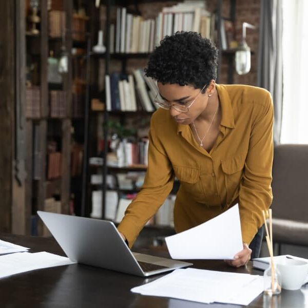 A woman at a desk examining documents and data on a laptop