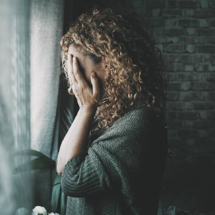 upset-woman-covering-her-face-with-her-hands-while-standing-at-a-window