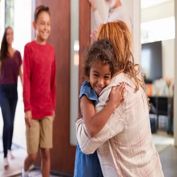 Little girl hugging her mother with her smiling siblings coming through the door behind her